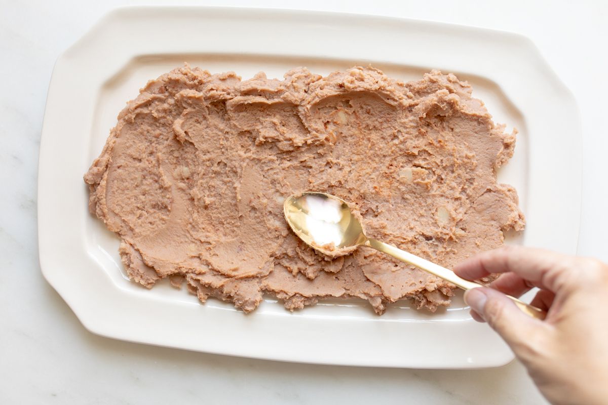 A spoon spreading refried beans onto a white platter for 7 layer taco dip.