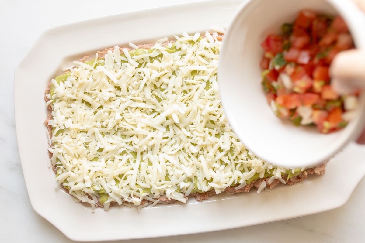 A white platter with refried beans topped with shredded cheese, a bowl of pico de gallo about to be spread across the top.