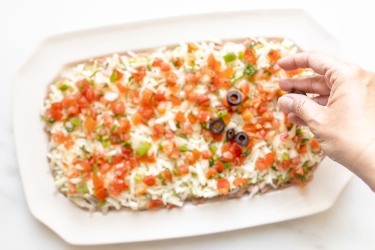 A hand adding garnish to a white platter of 7 layer dip.