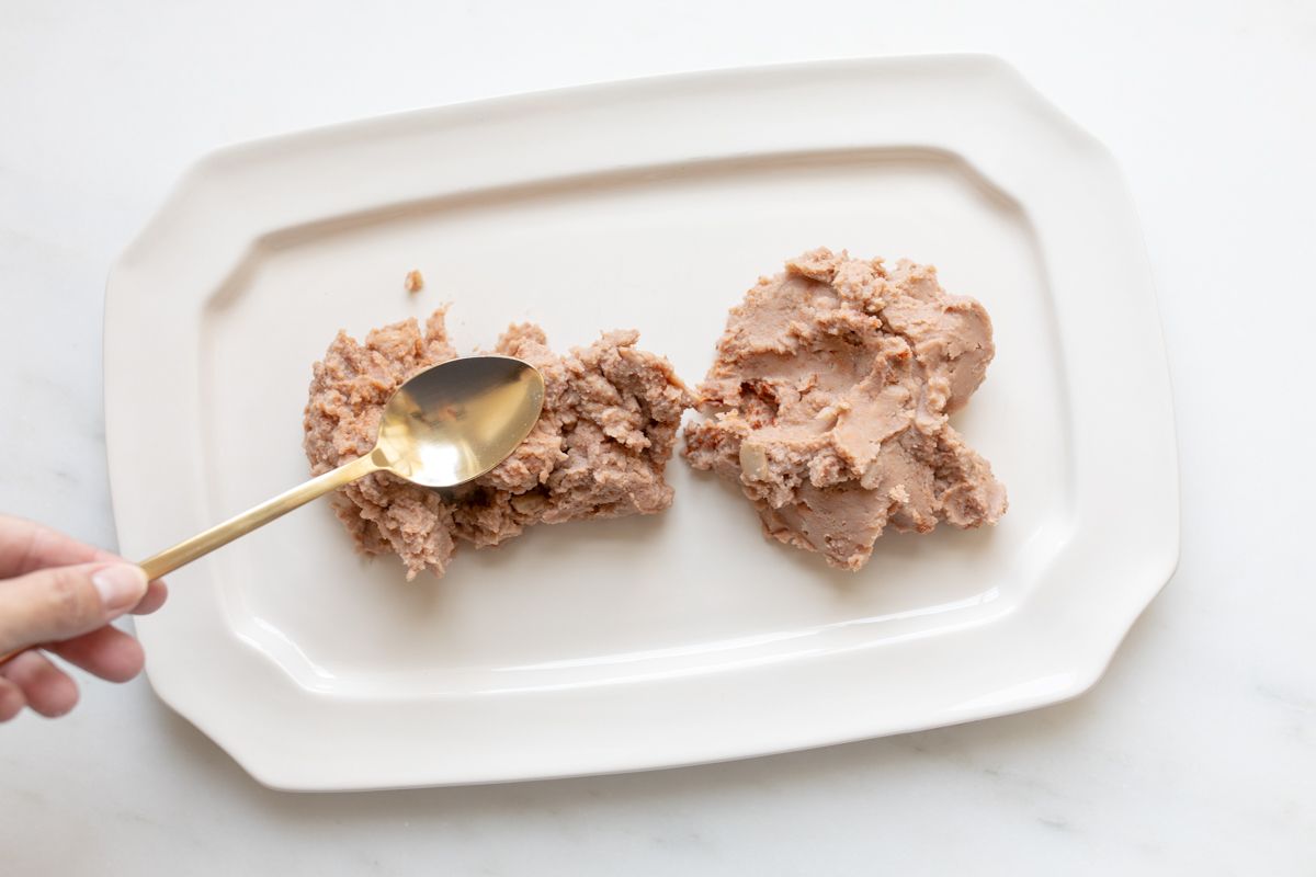 A spoon spreading refried beans onto a white platter for 7 layer taco dip.