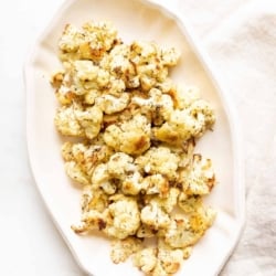 An oval white platter, filled with roasted cauliflower that is crispy and browned at the edges.