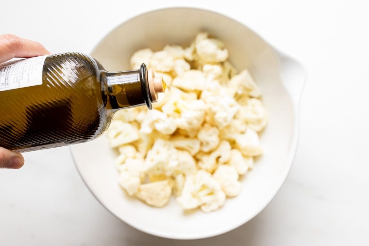 A bottle of olive oil being poured over a bowl of cauliflower florets.