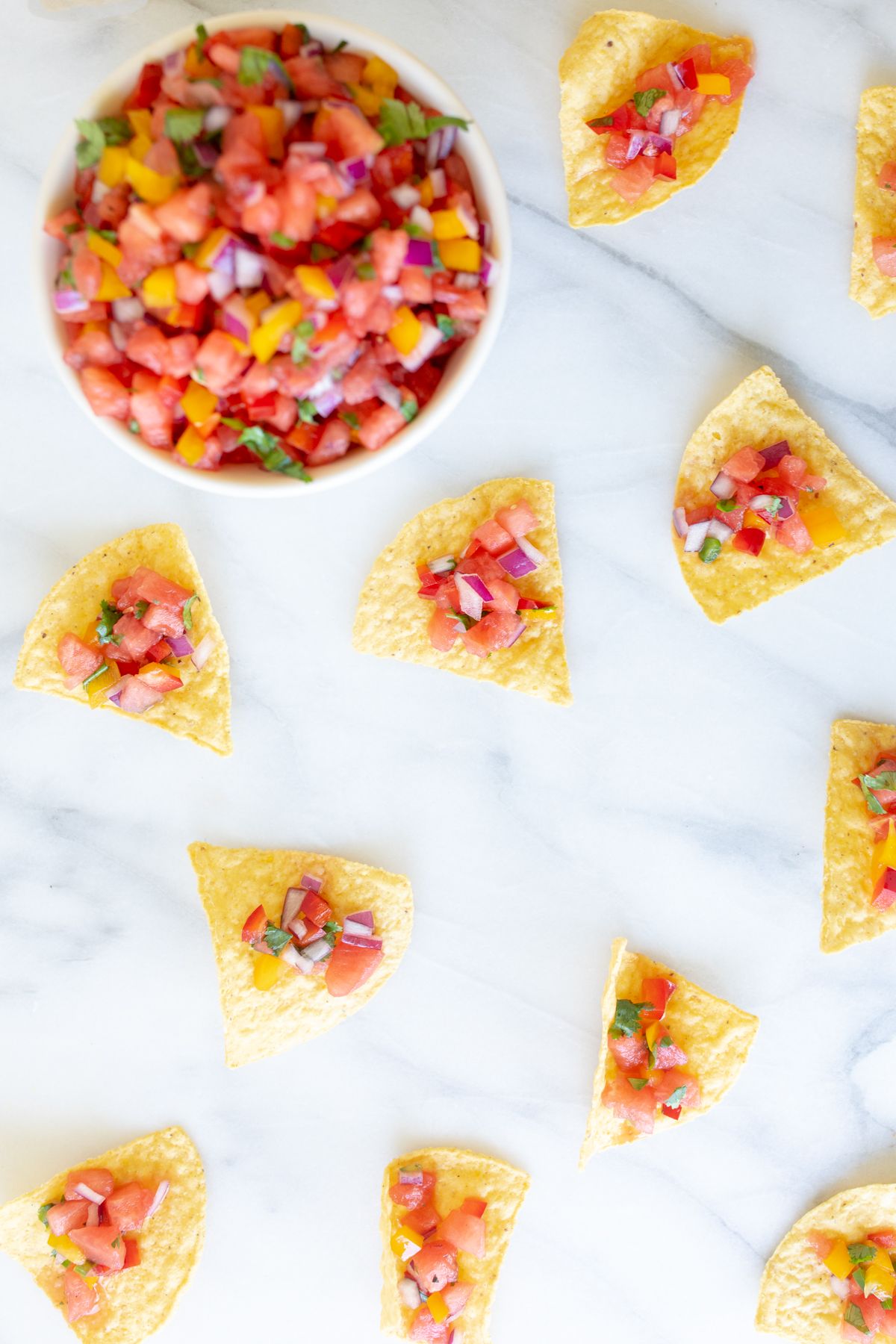 A marble countertop with tortilla chips scattered across, topped with fresh watermelon salsa. Bowl of watermelon salsa in the corner.