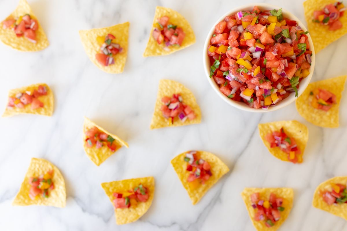 A marble countertop with tortilla chips scattered across, topped with fresh watermelon salsa. Bowl of watermelon salsa in the corner.