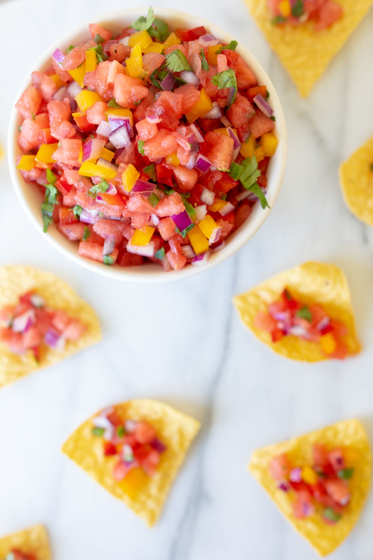 A marble countertop with tortilla chips scattered across, topped with fresh watermelon salsa. Bowl of watermelon salsa in the corner.