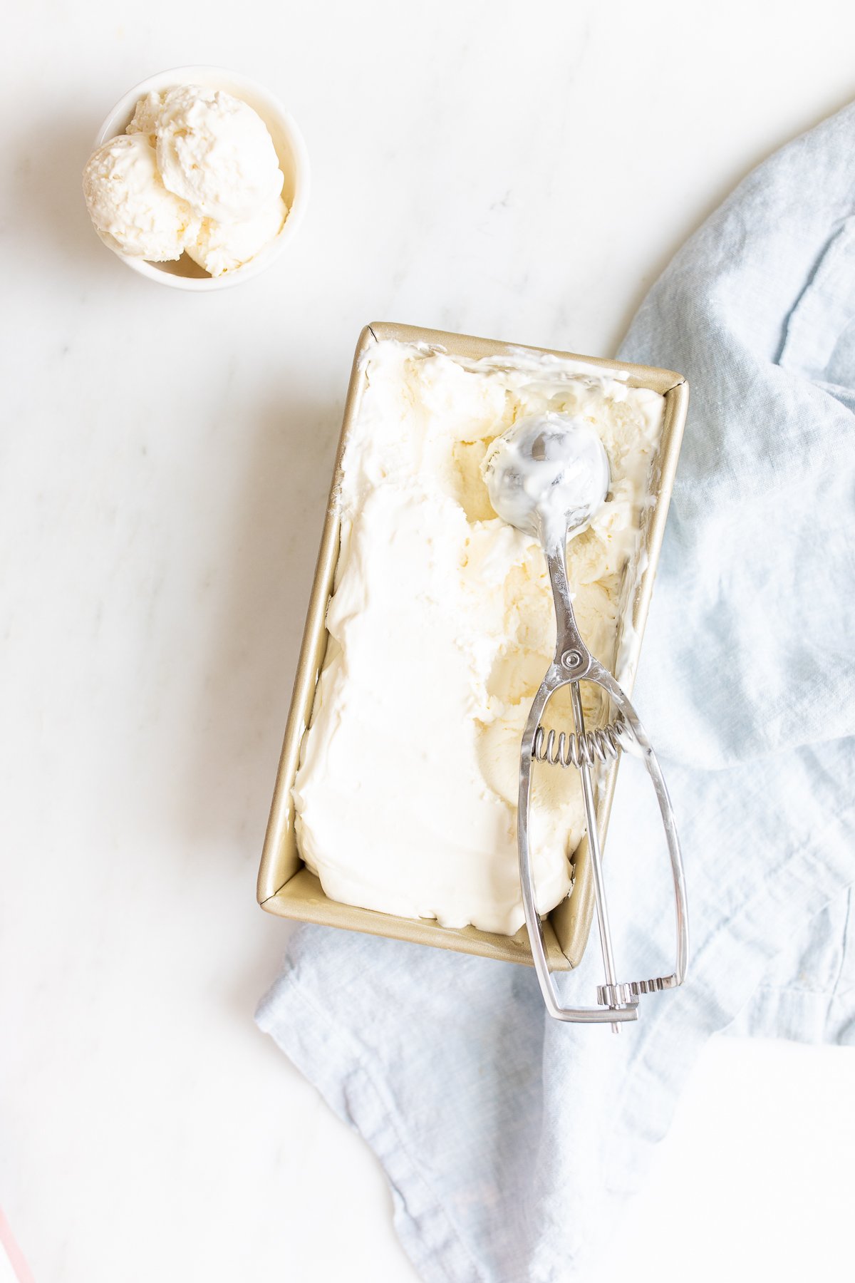 cream cheese ice cream in a loaf pan with an ice cream scooper and small white bowl of ice cream