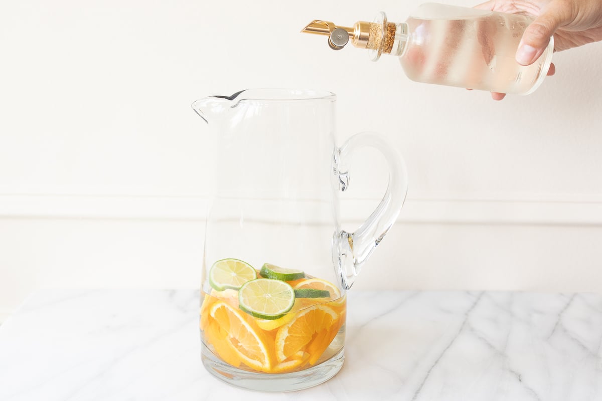 A glass pitcher full of fruit for red sangria, simple syrup being poured over the top.