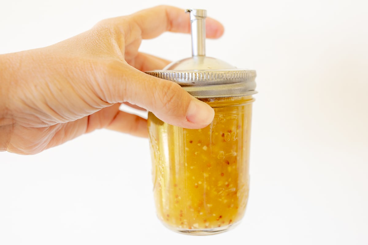 A hand holding a glass jar with a spout lid, full of pasta salad dressing