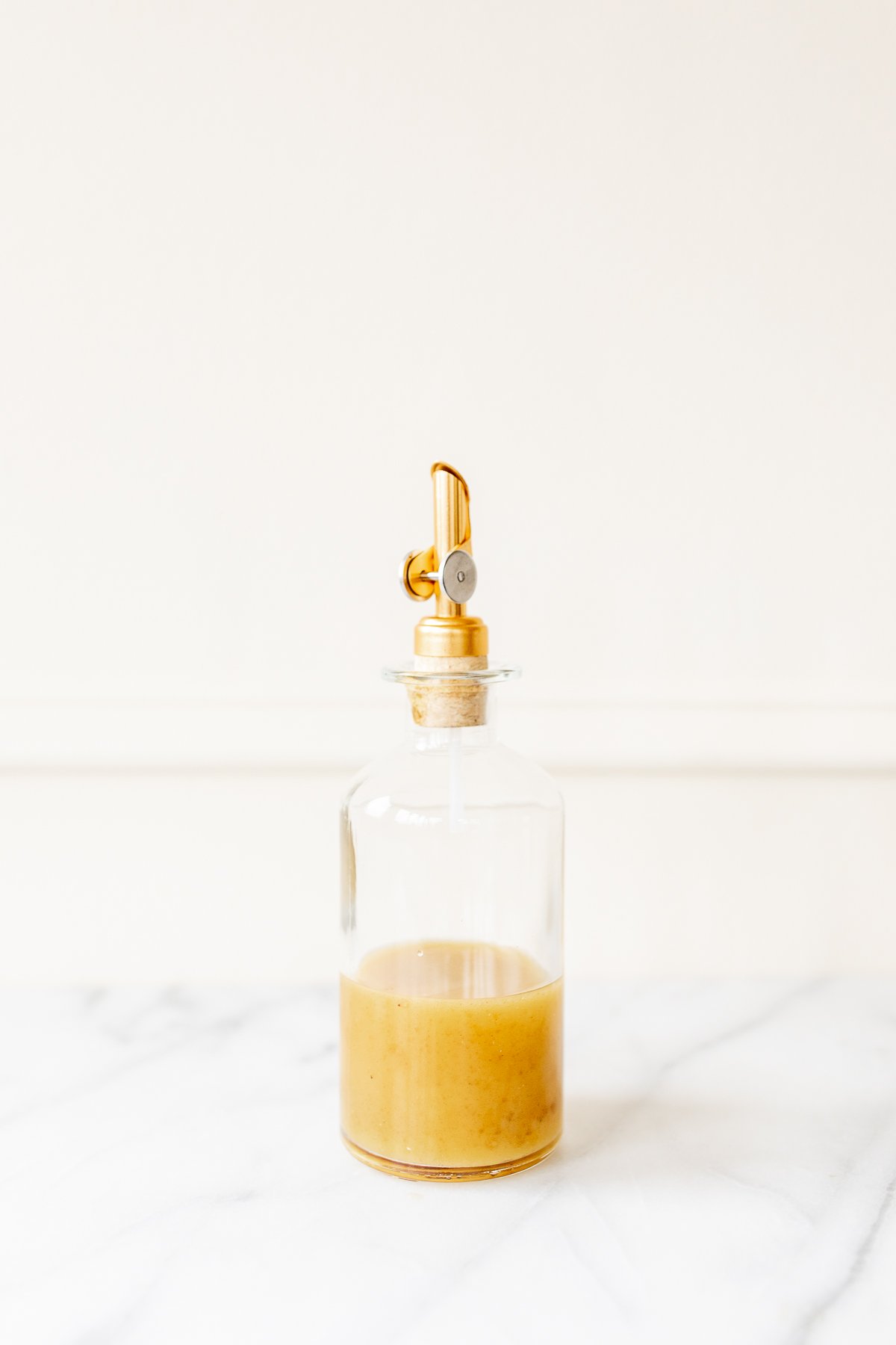 A glass bottle with a gold stopper on a marble countertop, filled with sherry vinaigrette.