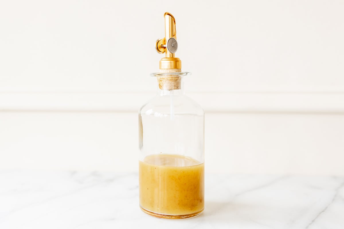 A glass bottle with a gold stopper on a marble countertop, filled with sherry vinaigrette.