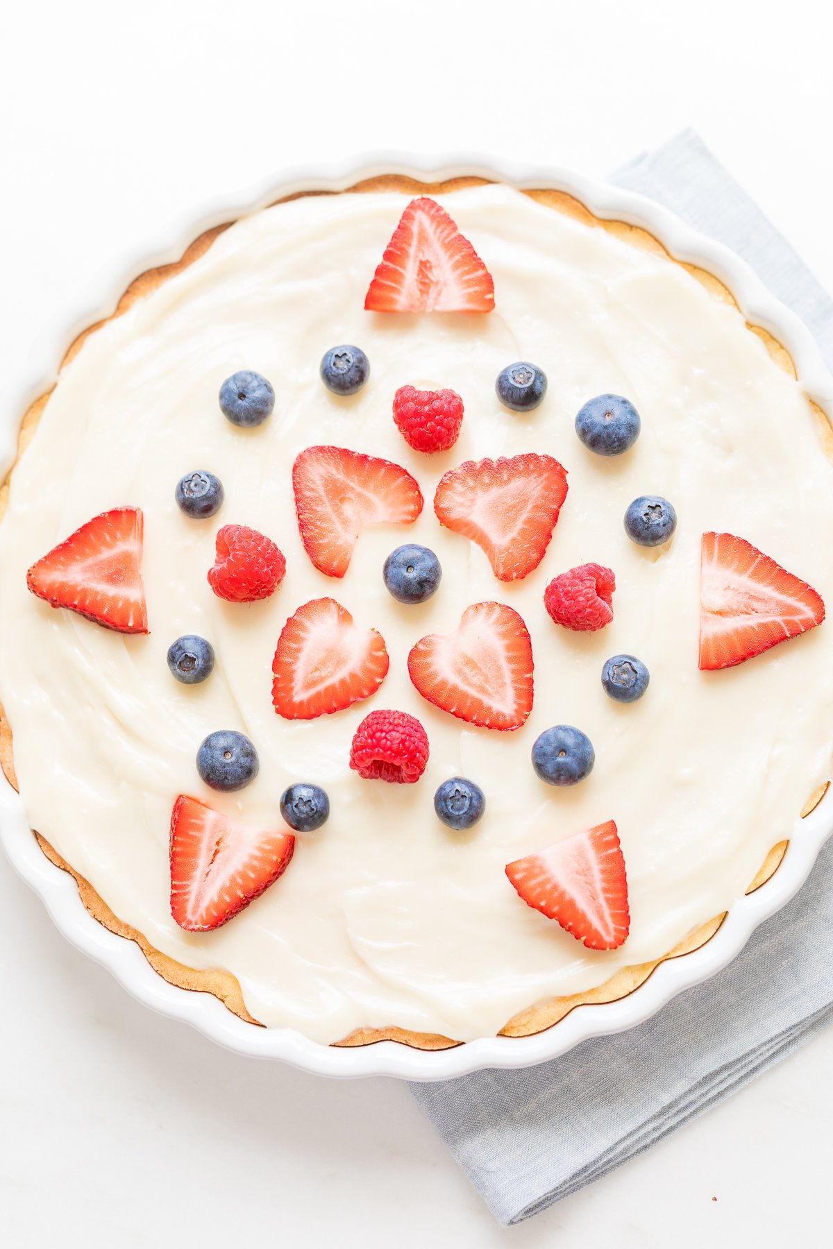 Fresh fruit tart with creamy filling and a fruit pizza glaze, garnished with sliced strawberries and whole blueberries on a white surface.