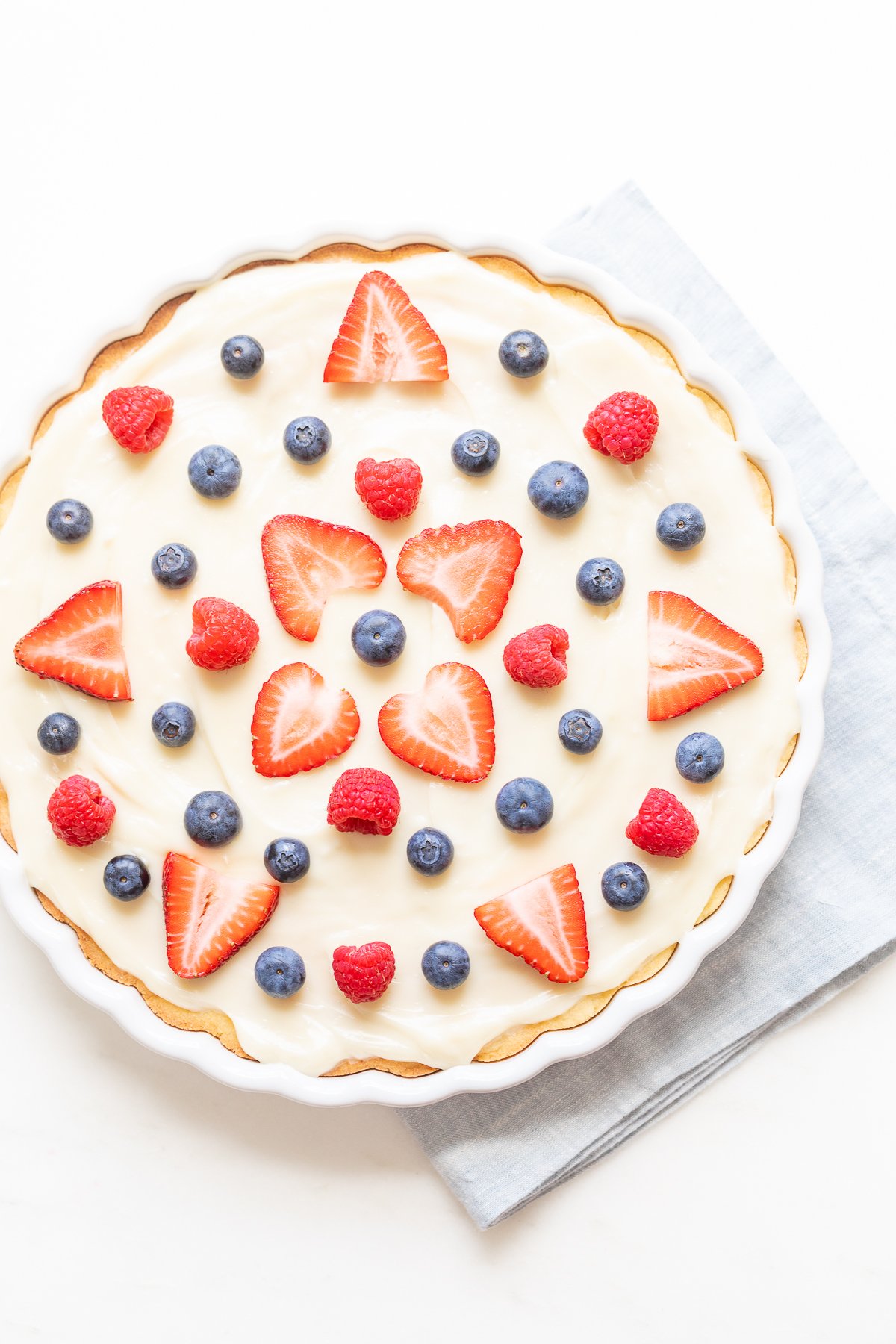 A fresh fruit pizza with cream and a decorative arrangement of strawberries, blueberries, and raspberries, finished with a glaze.