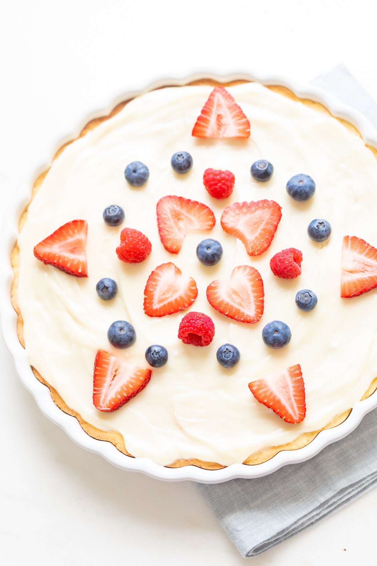 A fresh fruit pizza with cream topped with strawberries and blueberries on a white surface.