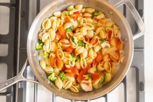 A stainless steel pan on a stove top with pasta shells and other ingredients to make pasta con broccoli.