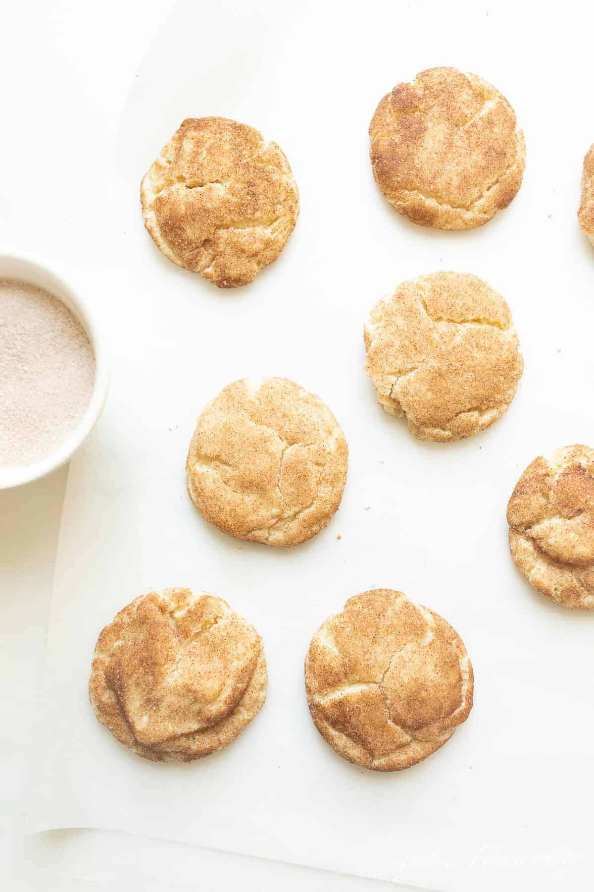 Snickerdoodle cookies on a white surface with a bowl of cinnamon sugar to the side. 