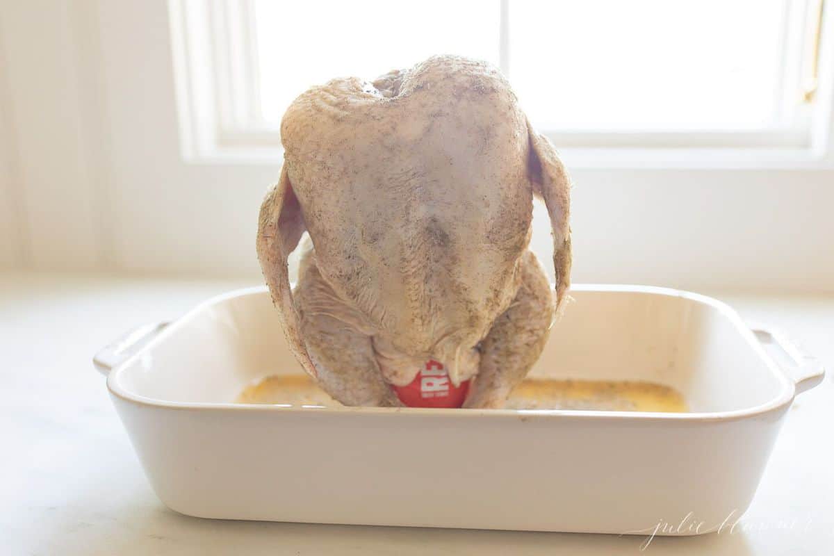 Raw whole chicken in a white roasting pan, resting on a beer can with beer at the base of the pan.