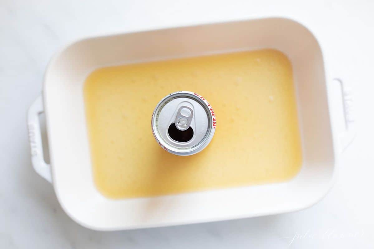 Can of beer inside a white roasting pan with beer at the bottom of the pan, on a white surface.
