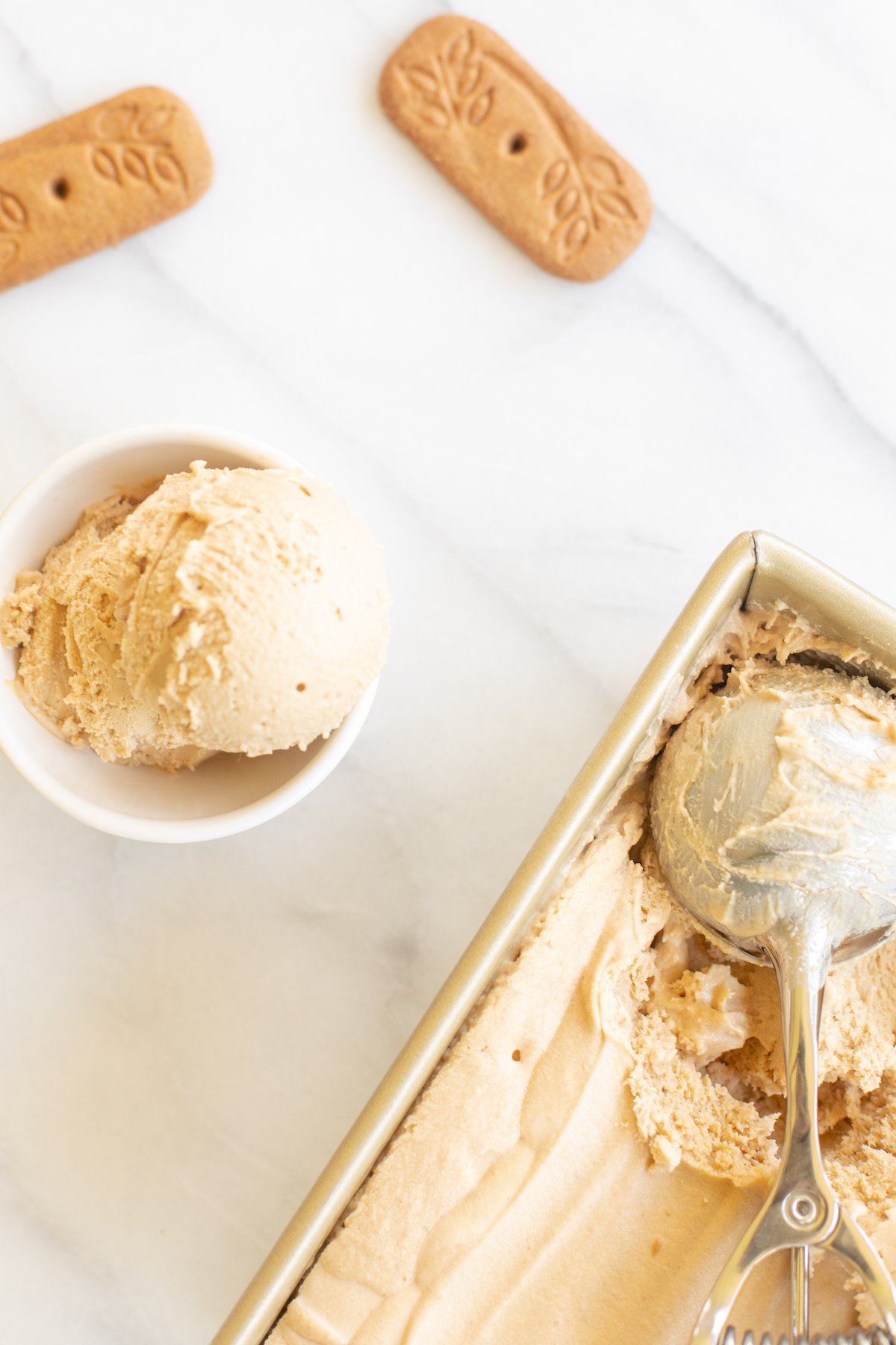 A gold loaf pan full of homemade no churn cookie butter ice cream. Bowl of ice cream and speculoos cookies to the side.