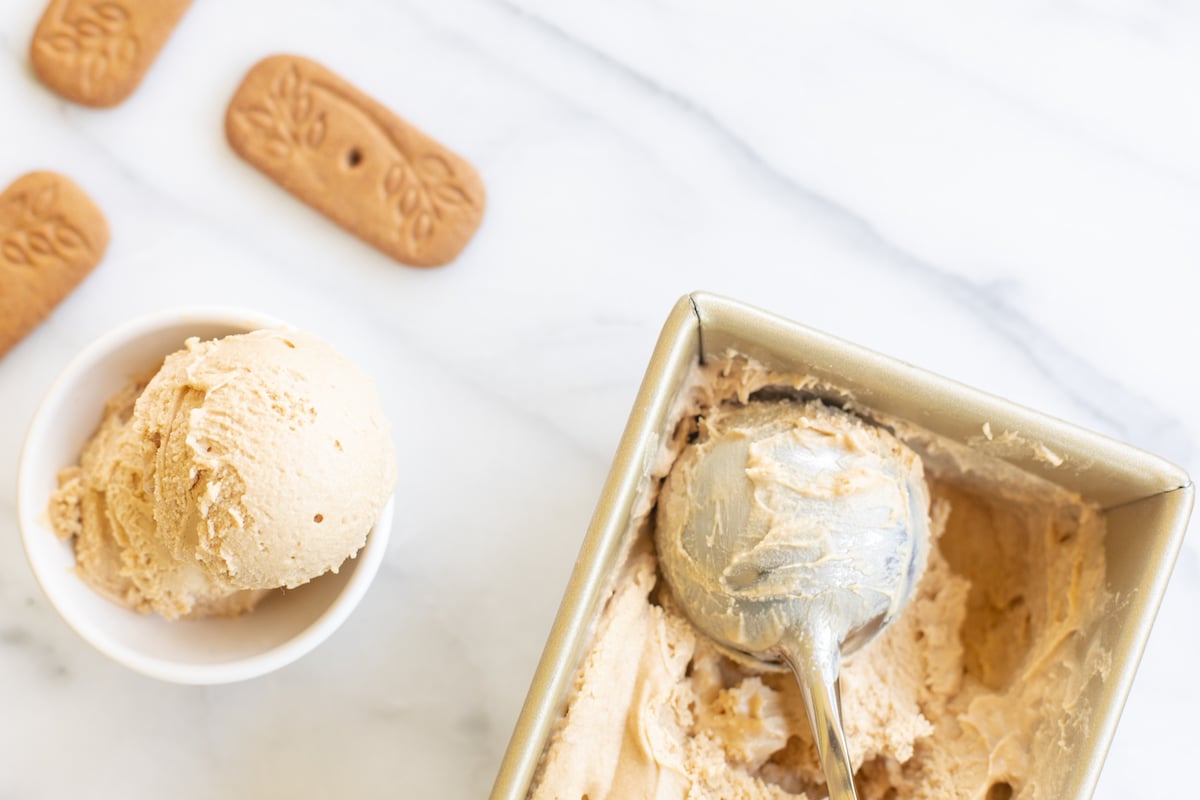 A gold loaf pan full of homemade no churn cookie butter ice cream. Bowl of ice cream and speculoos cookies to the side.