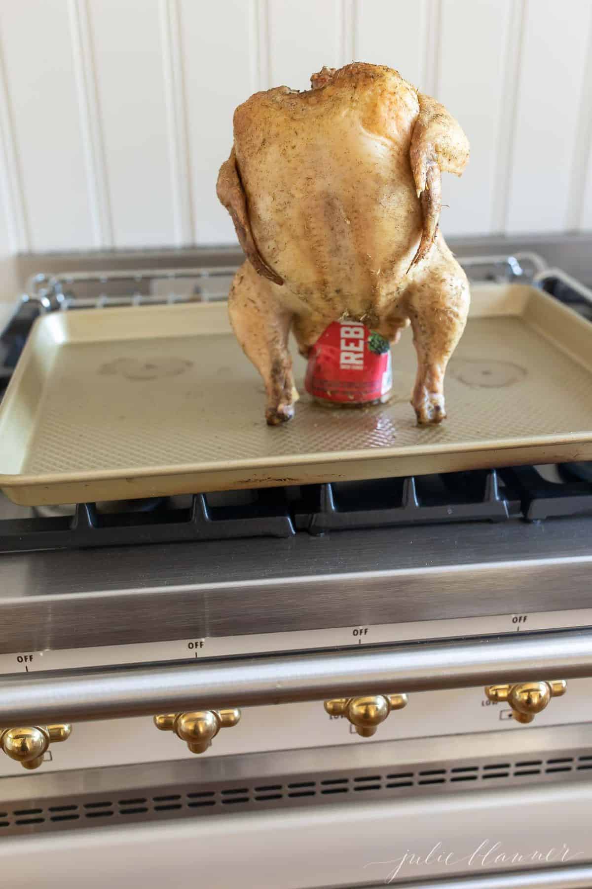 Verical image of roasted chicken resting on a beer can, placed on a gold sheetpan on top of range. #beercanchicken
