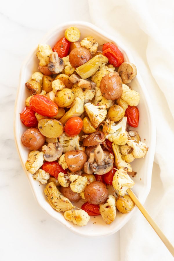 A dish of Italian roasted vegetables including potatoes, cauliflower, tomatoes, and mushrooms, served in a white oval platter on a marble surface.