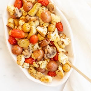 Italian roasted vegetables including potatoes, cauliflower, and tomatoes in a white serving dish on a marble countertop.