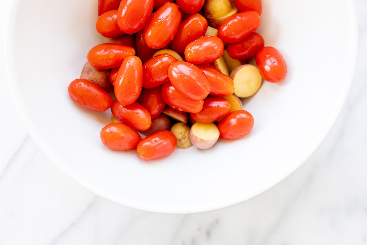 A white bowl containing red cherry tomatoes and yellow potatoes on a marble surface, ready to be used in Italian vegetable dishes.