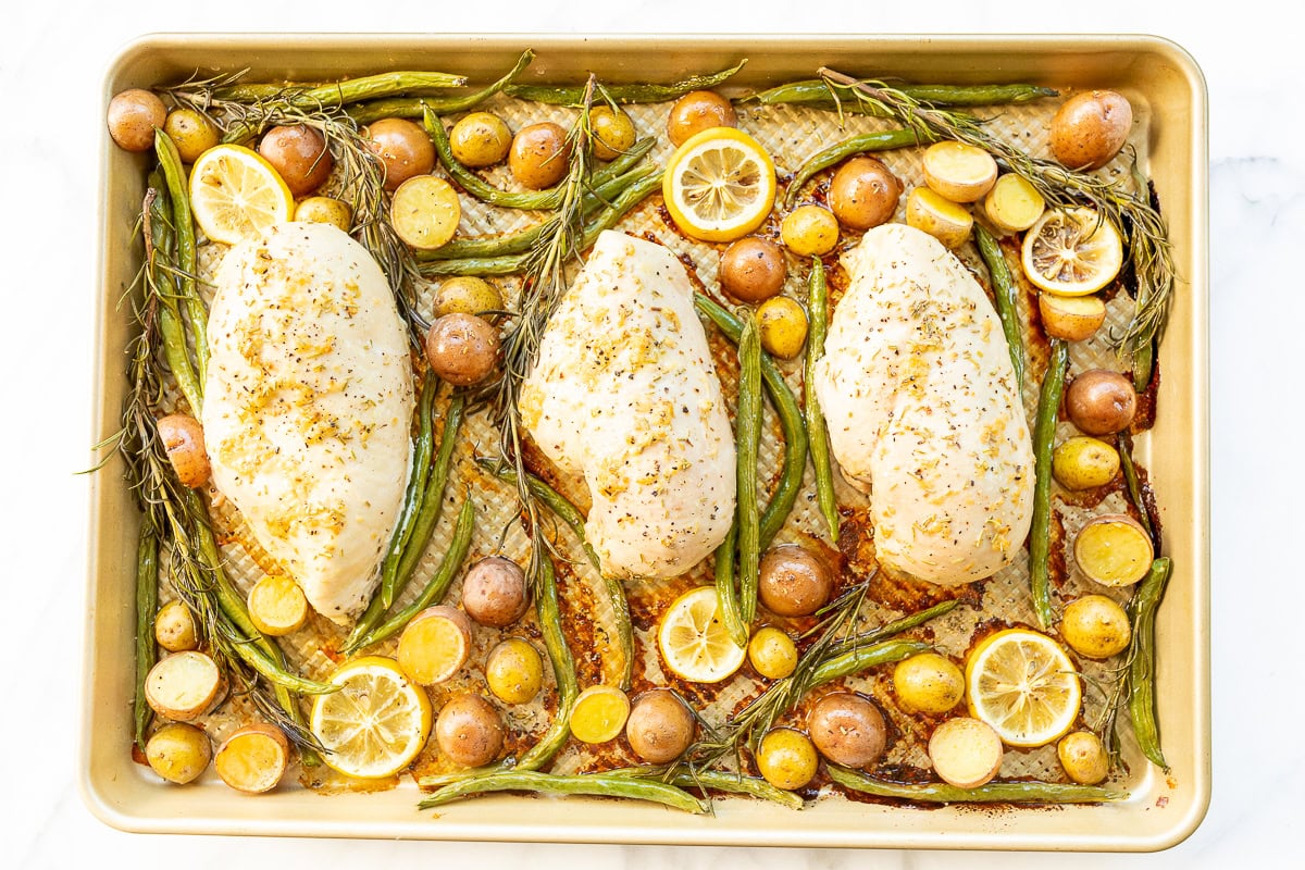 Three rosemary chicken breasts with lemon slices, green beans, and potatoes on a baking sheet, seasoned with spices.