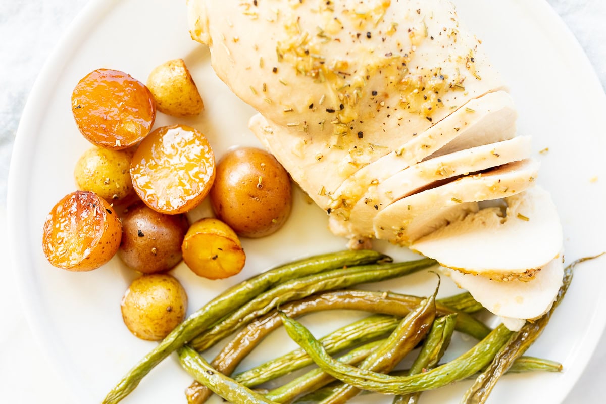 A plate with sliced rosemary roasted chicken breast, roasted potatoes, green beans, and halved cherry tomatoes.