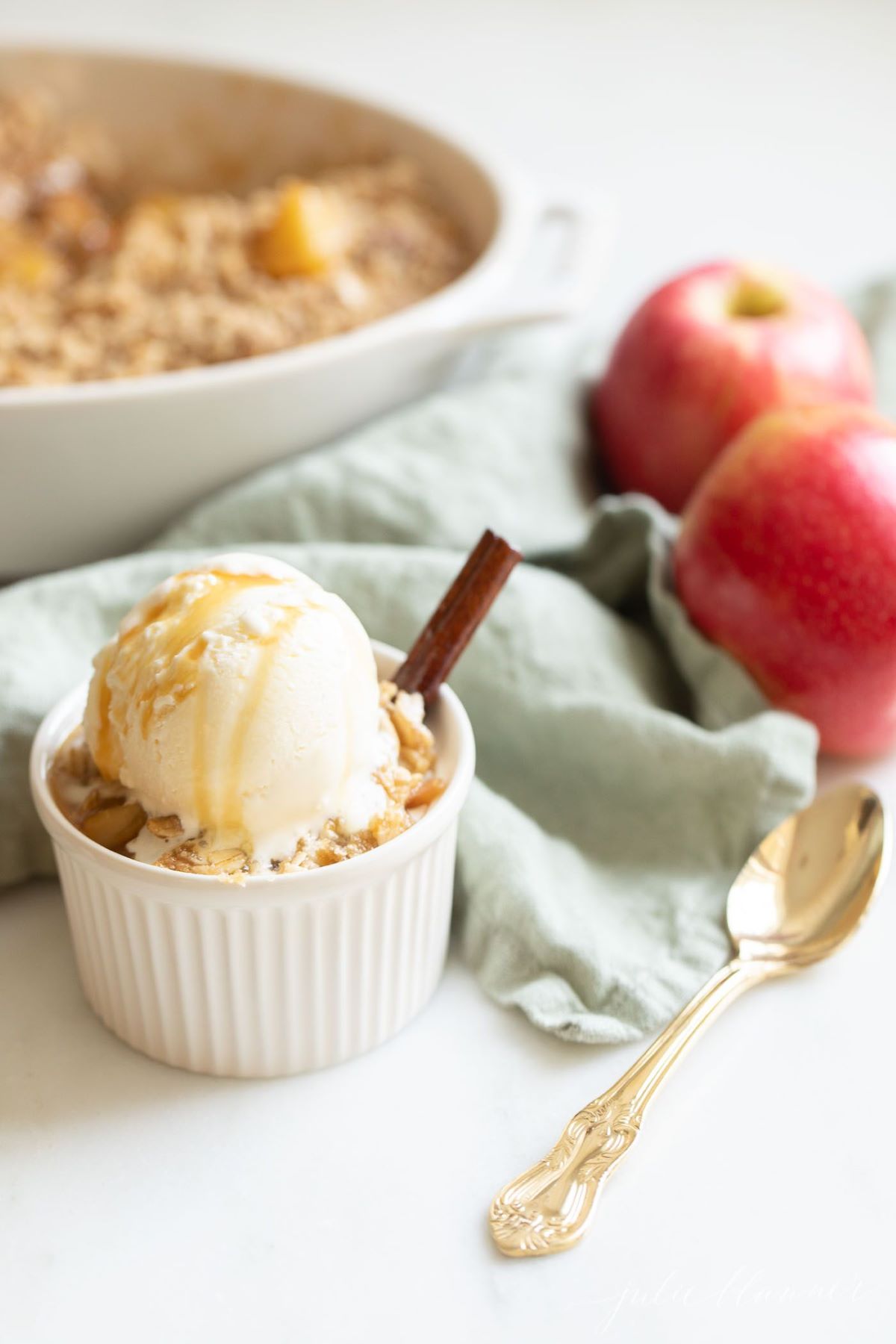 Apple crisp topped with ice cream in a small white ramekin, apples and the whole pan of crisp in the background