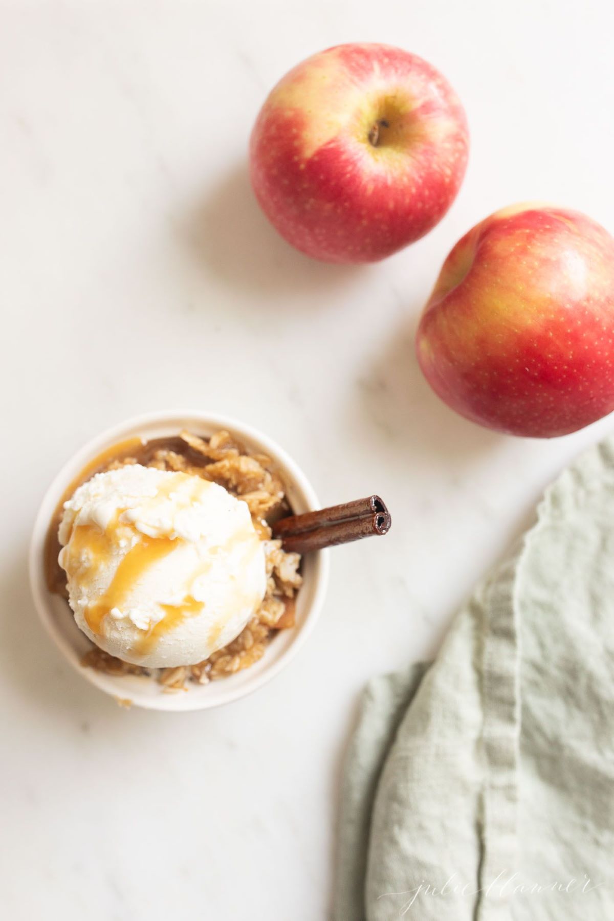 Apple crisp topped with ice cream in a small white ramekin, apples in the background