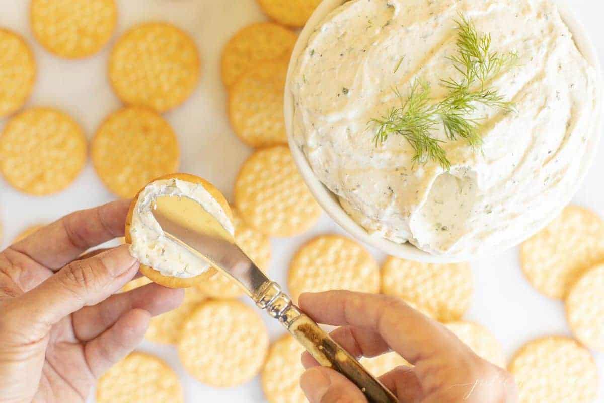 White bowl of herb cream cheese with crackers, and a hand spreading cheese onto a cracker. #homemadeboursin