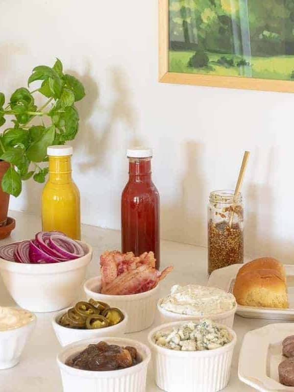 Burger toppings displayed in white ceramic bowls, condiments in rear and buns to the side, burgers in foreground. #burgerbar #burgershop #sliderbar