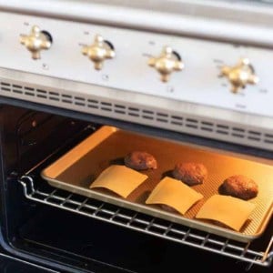 Looking into a range with the door open, burgers under broiler on a baking sheet, half of them have cheese.