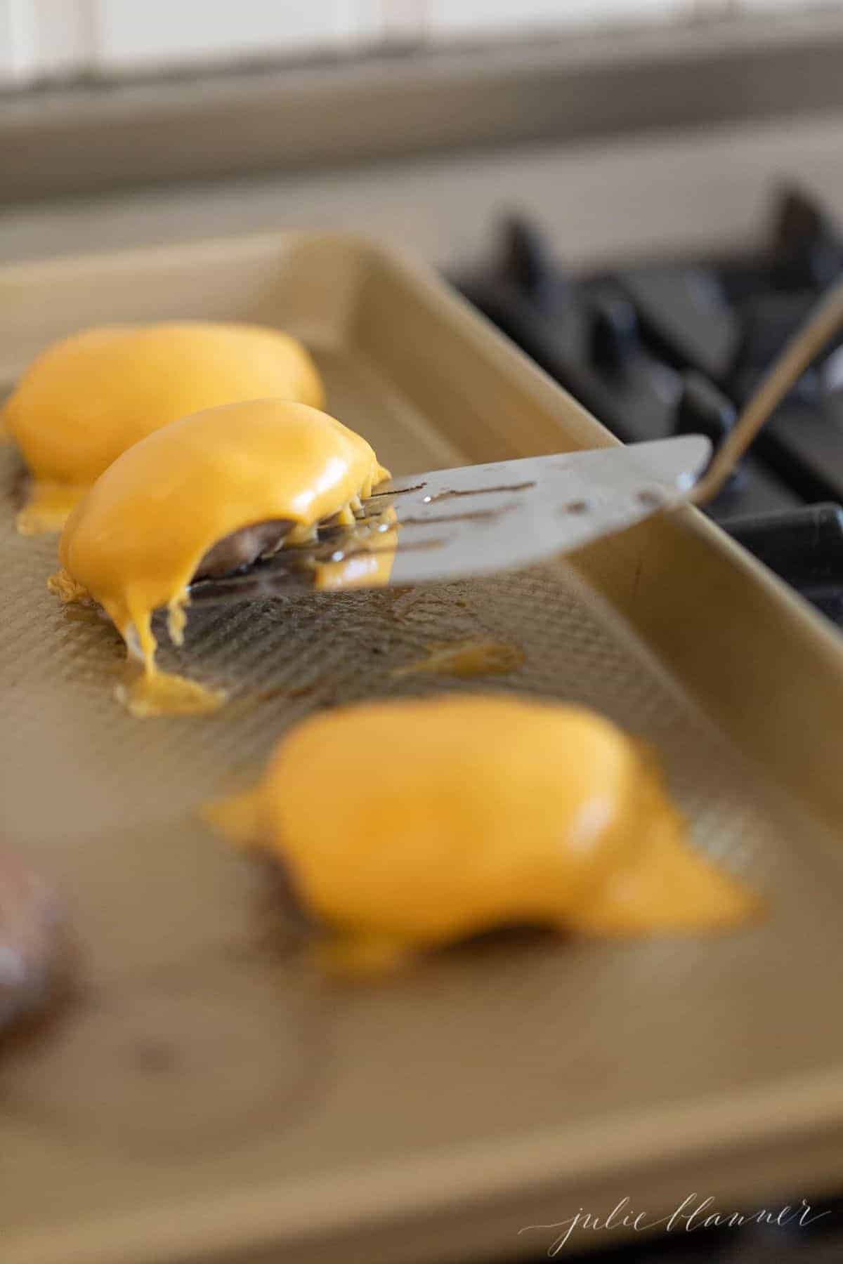 Burger patties covered in cheese, raising off a baking sheet with a metal spatula. 