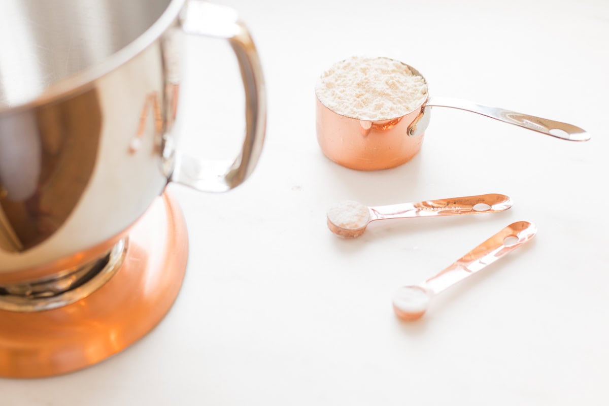 Copper measuring cups and spoons filled with flour are placed on a white surface next to a metallic mixing bowl, accompanied by a handy measurement conversion chart.