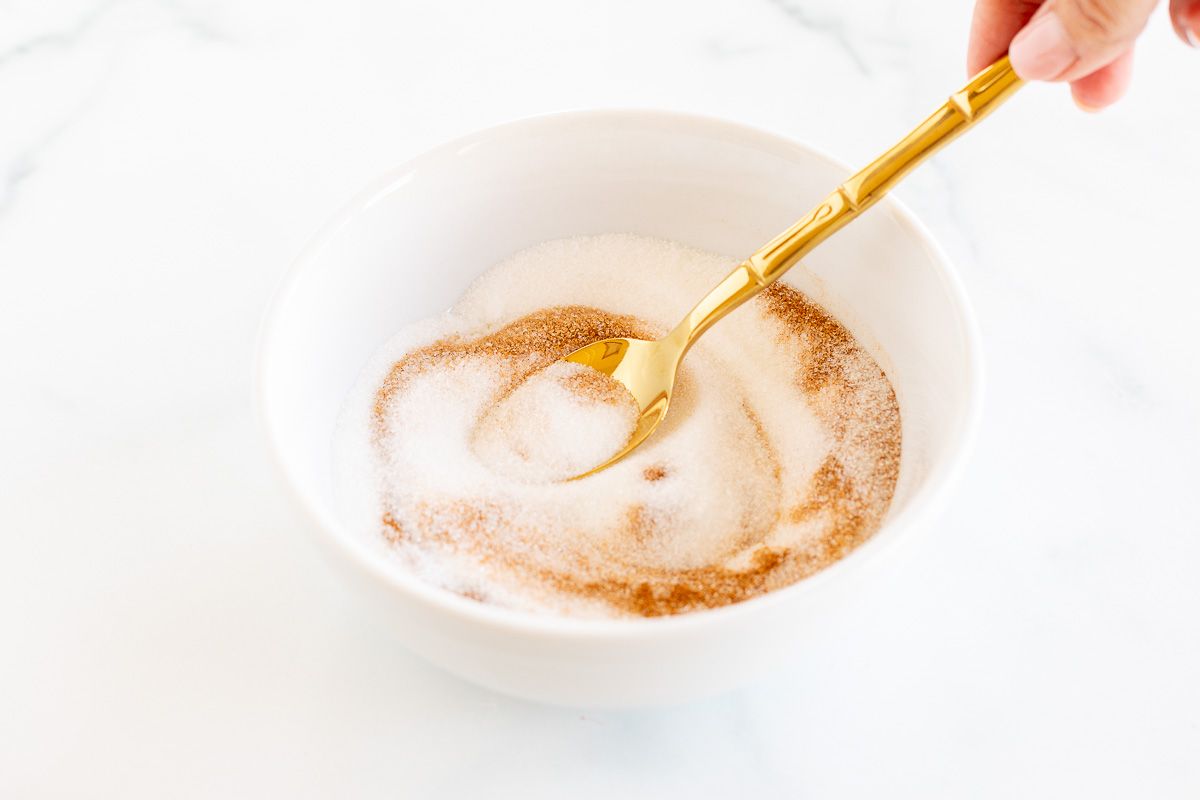 A bowl of cinnamon sugar with a hand stirring a gold spoon.