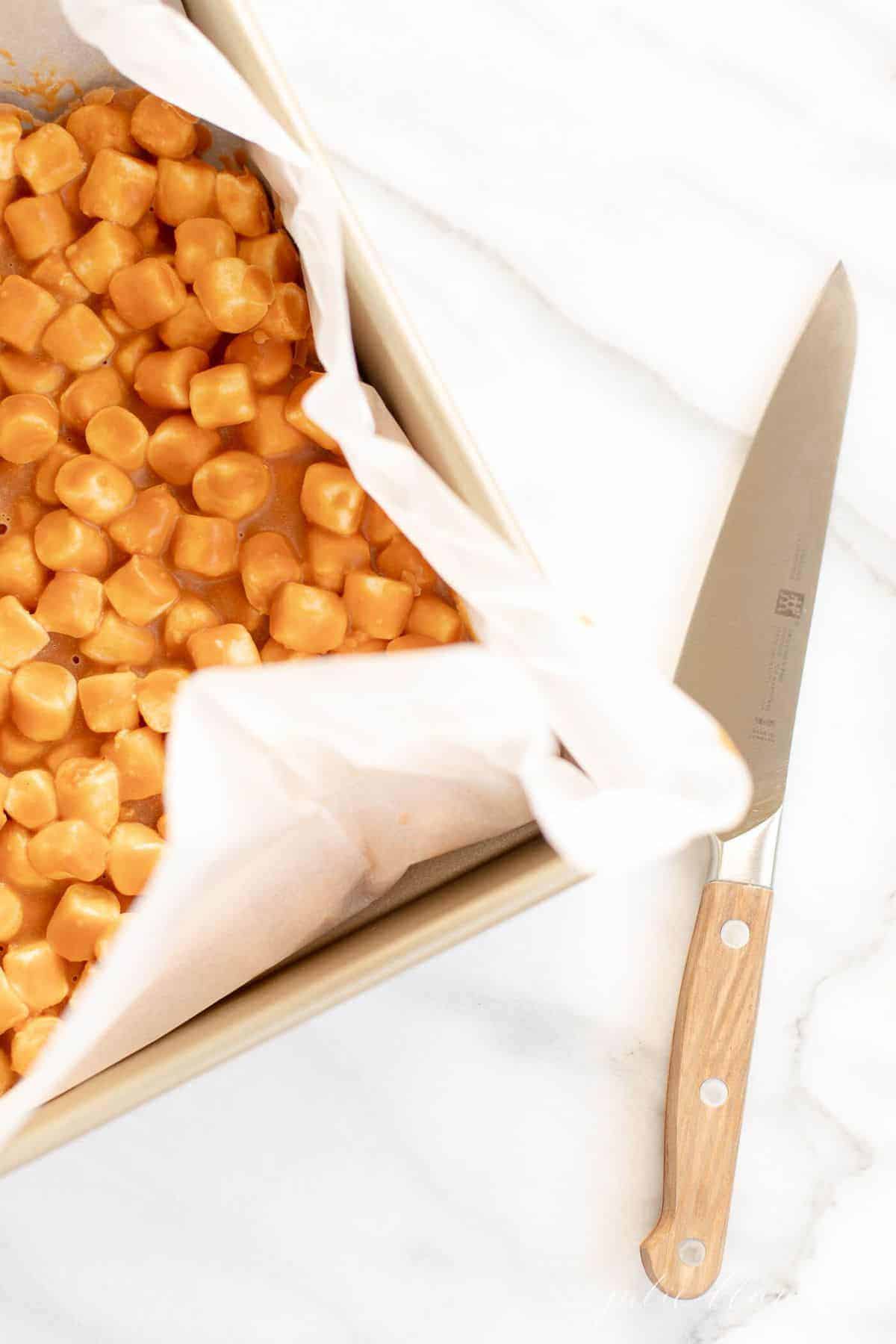 Gold pan with parchment paper lining, butterscotch squares recipe inside and knife to the side.