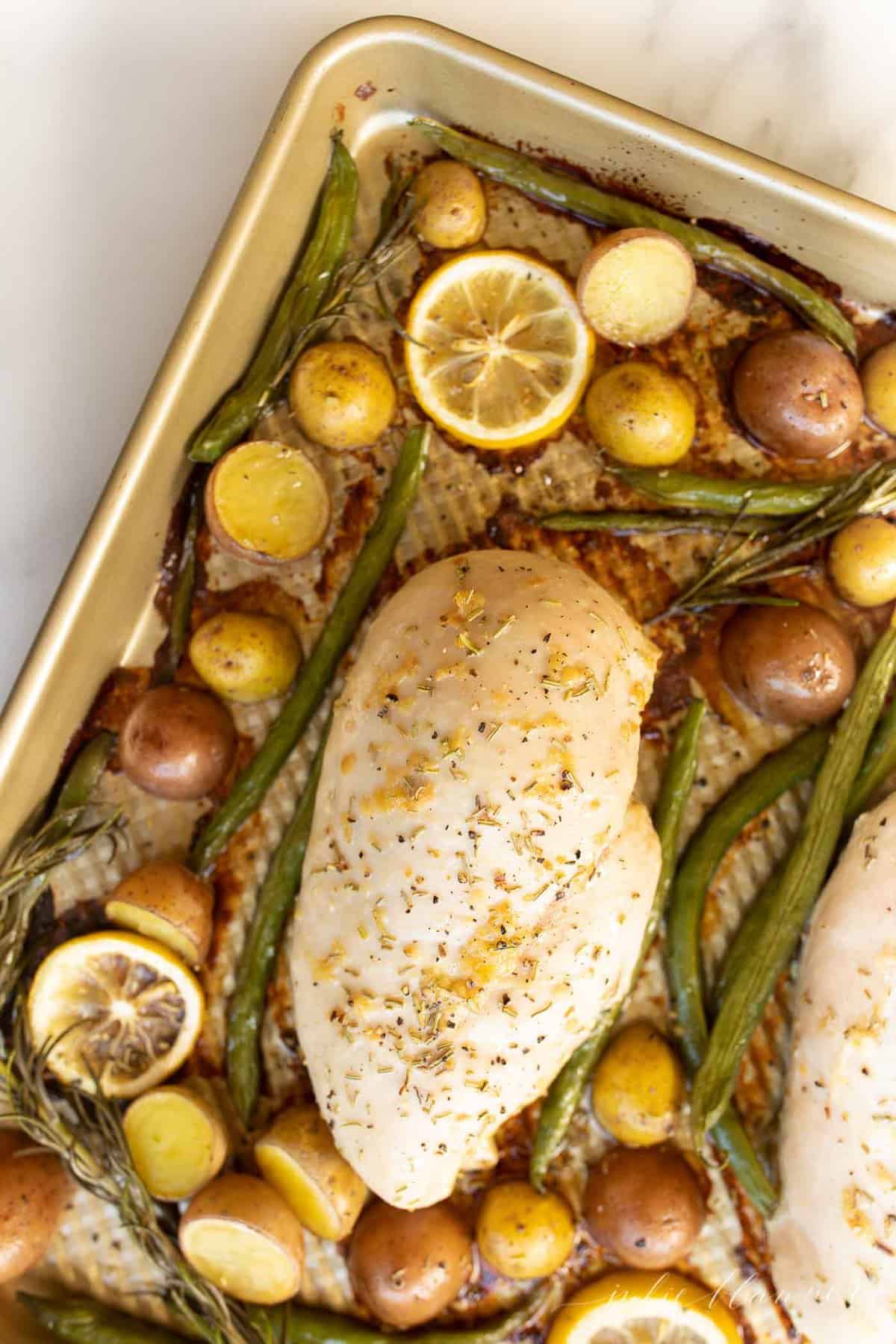 Gold baking sheet with a baked chicken breast, lemon slices and roasted veggies.