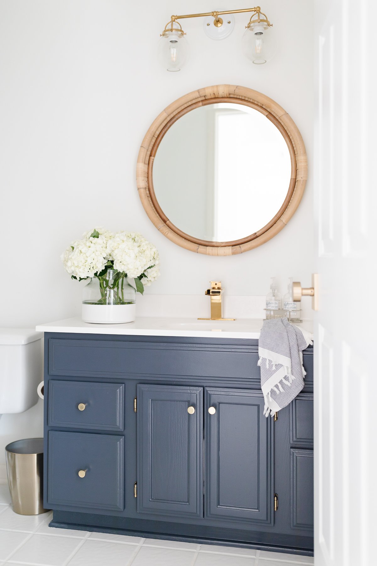 A nautical bathroom with a navy vanity and a rattan round mirror.