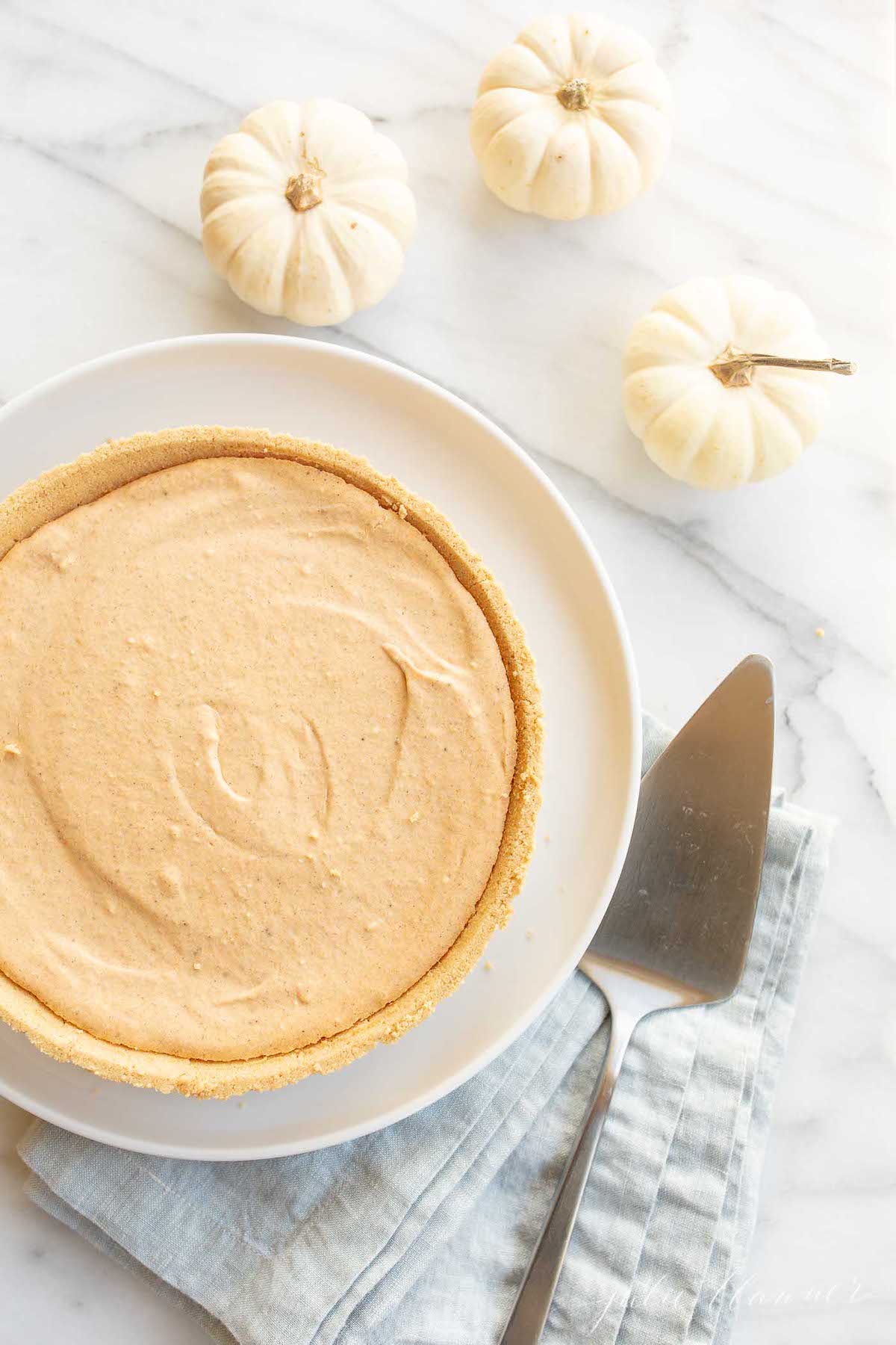 A no bake pumpkin cheesecake on a white plate, with a pie server to the side and small white pumpkins in the background.