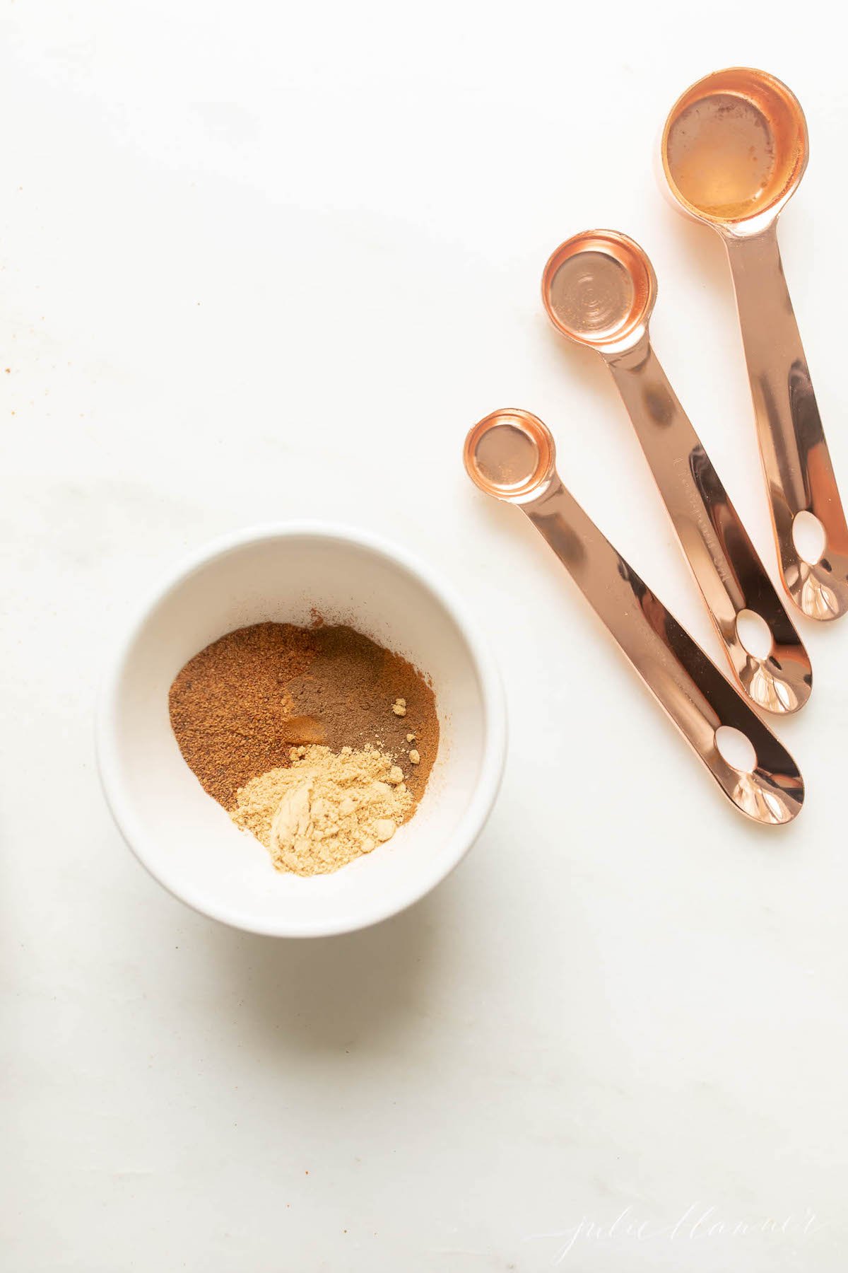 A white bowl full of spices for a pumpkin pie spice recipe. Copper measuring spoons to the side.
