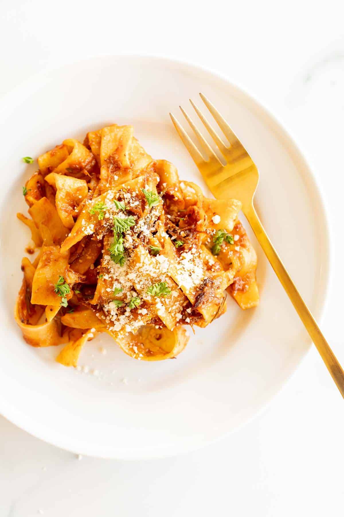 A small white plate with beef ragu and pappardelle pasta, gold fork to the side.