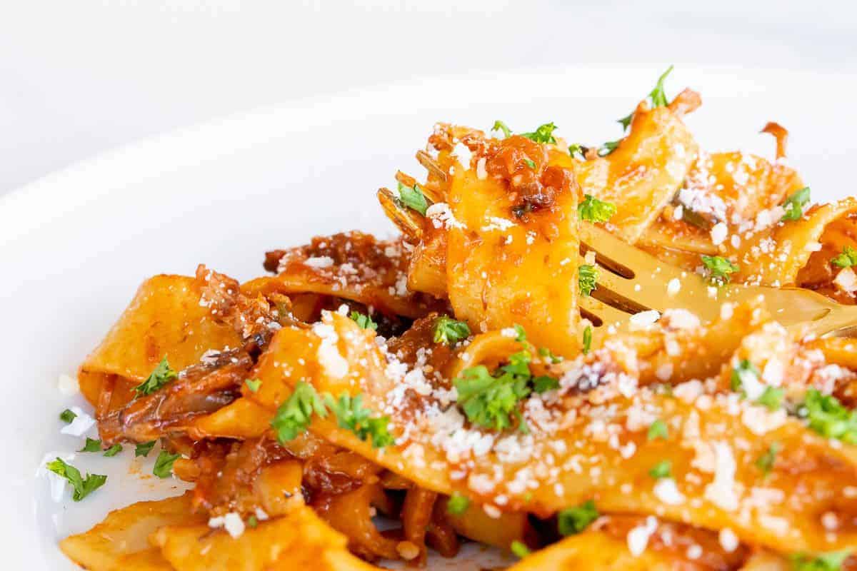 A small white plate with beef ragu and pappardelle pasta, gold fork to the side.