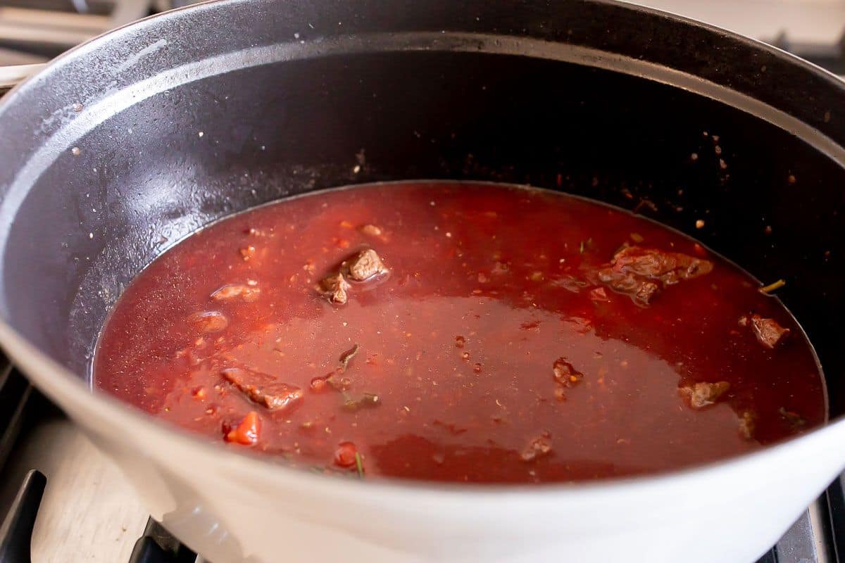 A white cast iron pot on a stove, filled with beef ragu sauce simmering.