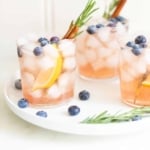Three glasses of bourbon smash cocktails on a white tray, garnished with blueberries, lemon, rosemary and cinnamon
