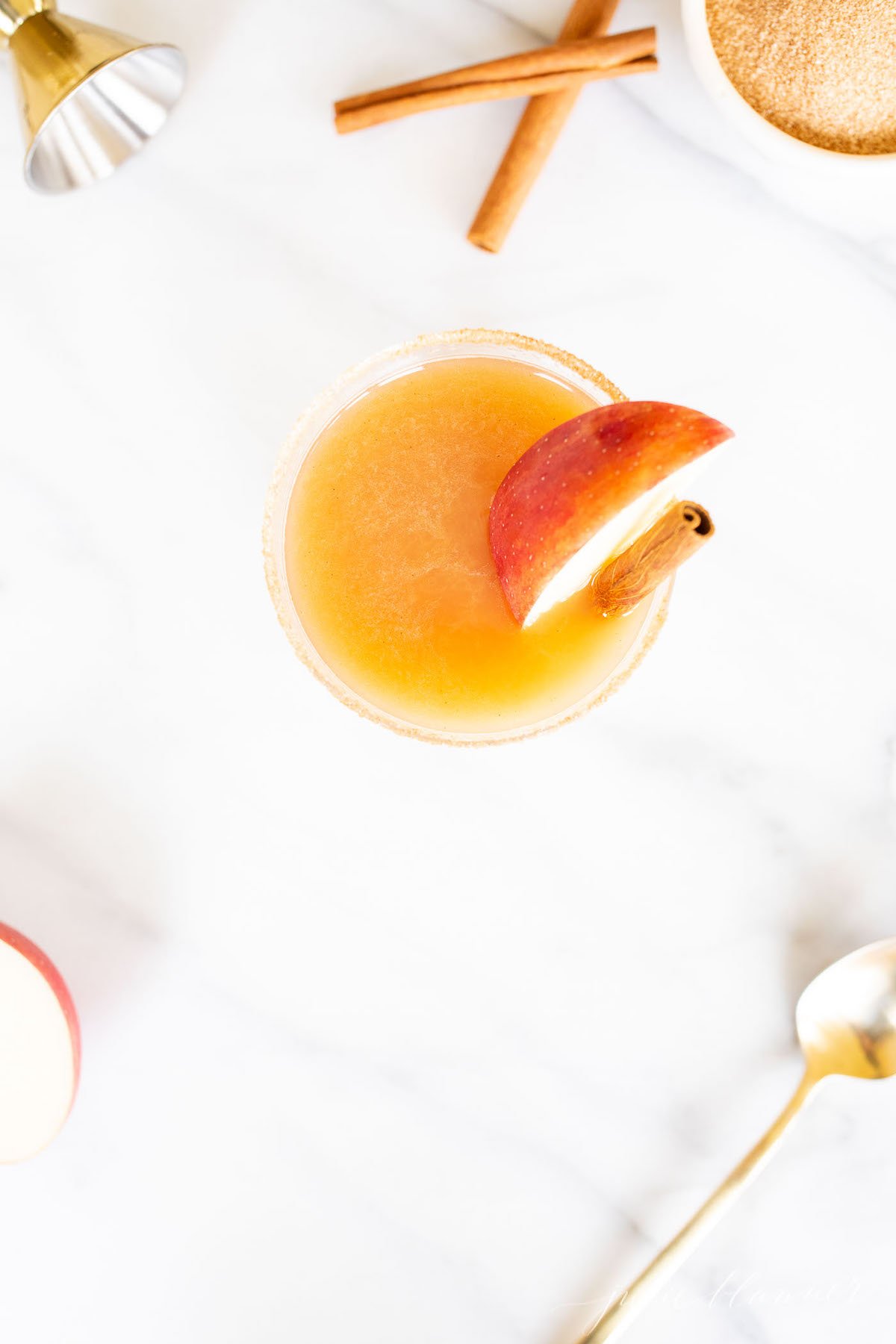 Cinnamon liquor cocktail glass on marble surface, garnished with apple slice and cinnamon sticks.