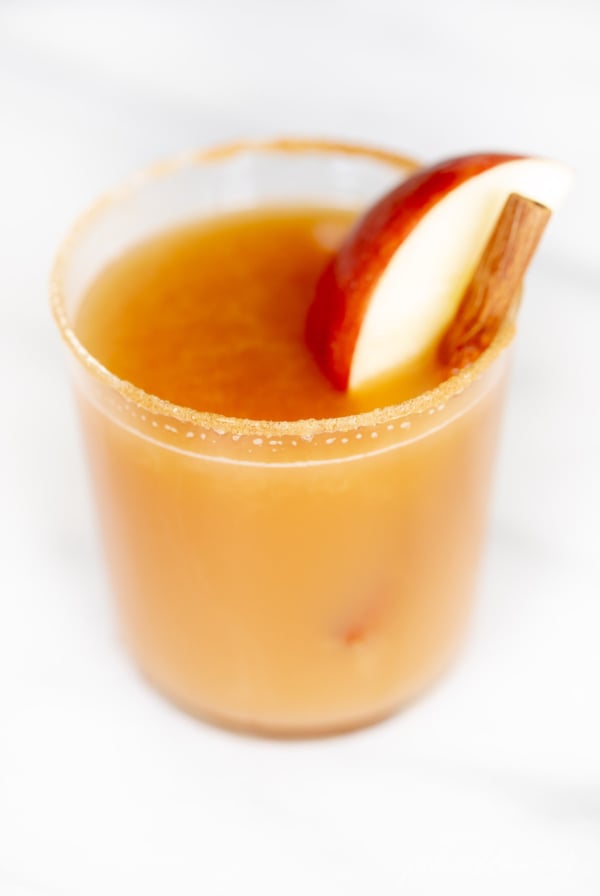 Cinnamon toast cocktail in a glass on marble surface, garnished with apple slice and cinnamon sticks.