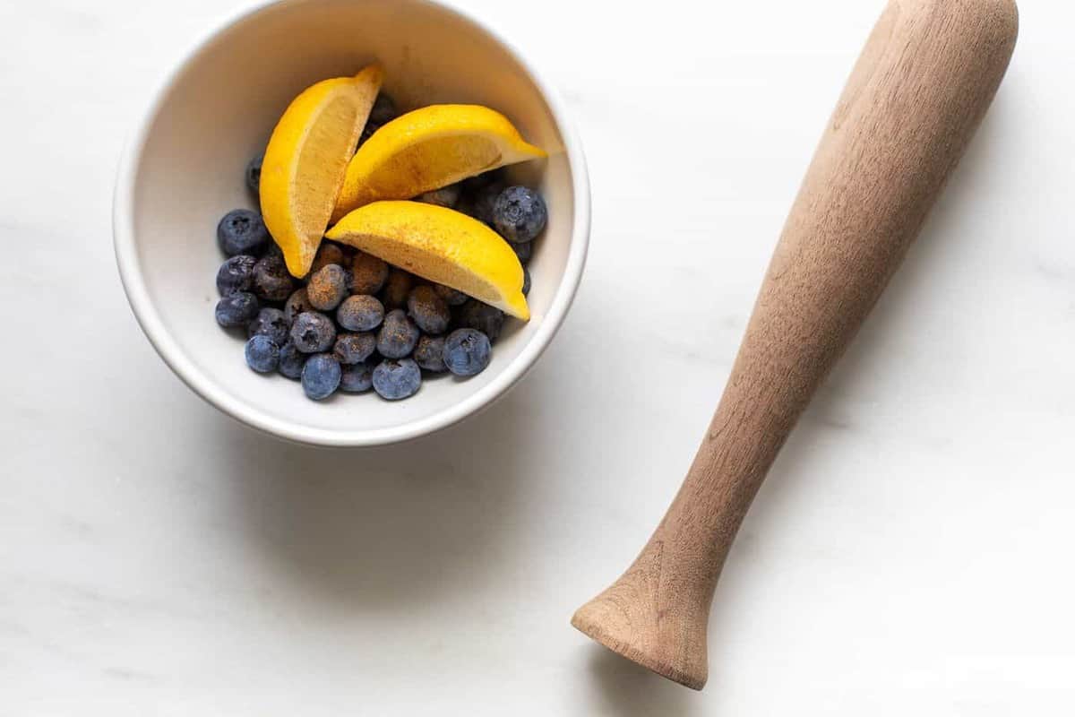 marble surface, bowl of lemon slices and blueberries with a wooden muddler.
