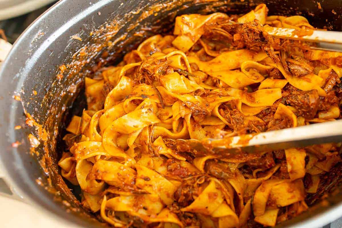 Beef ragu and Pappardelle pasta in a large cast iron pot, wooden spoon to the side.